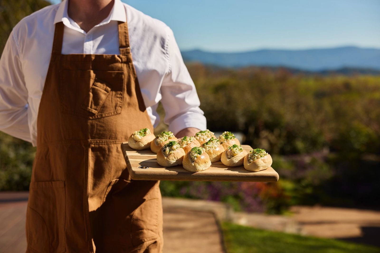 A Greyleigh team member in a leather apron carrying a board of canap&eacute;s, hills behind