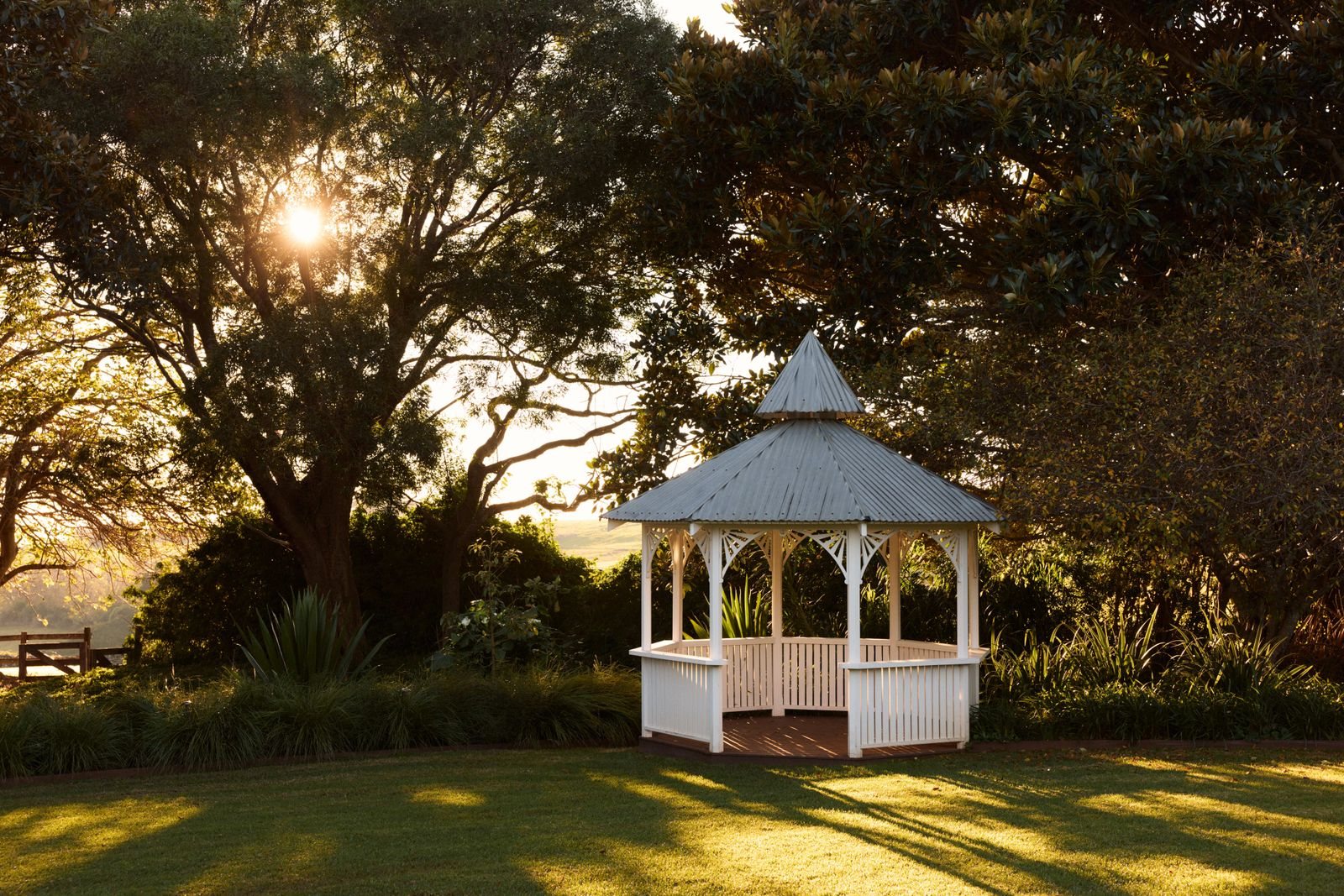 The white gazebo on the lawn under old trees at sunset