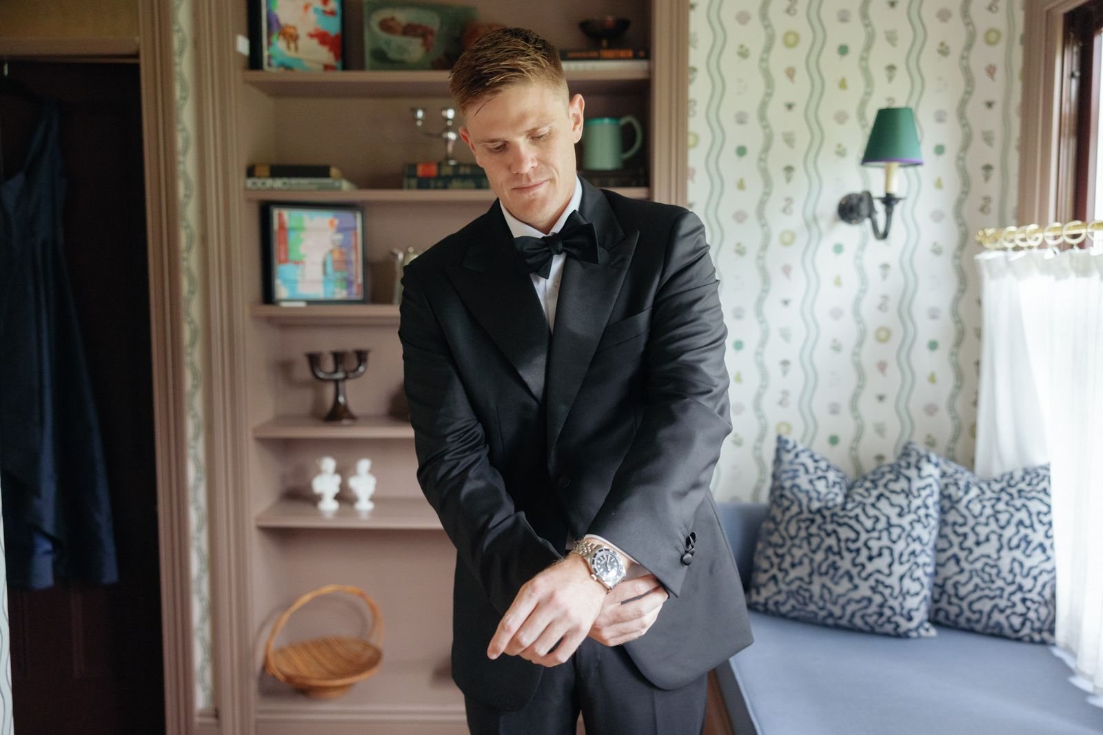 A groom in a dinner suit fixing his cuff in a patterned Homestead room